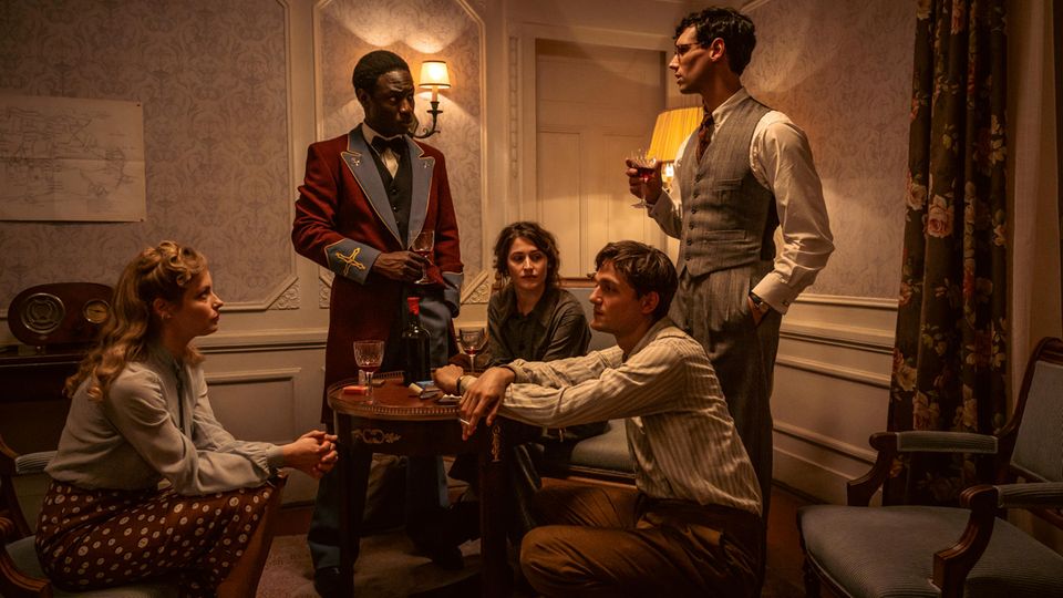 Filmszene aus "Transatlantic": Gruppe von Menschen berät sich auf einem Hotelzimmer