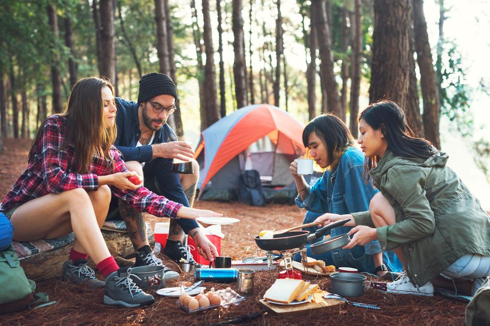 Eine Gruppe junger Leute sitzt um den Camping Kocher und bereitet gemeinsam Essen zu.