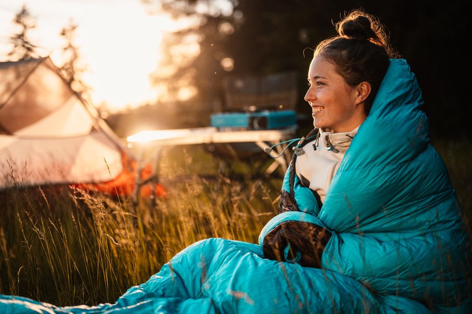 Eine junge Frau sitzt in einen Schlafsack eingewickelt in der Nähe eines Campingzelts.