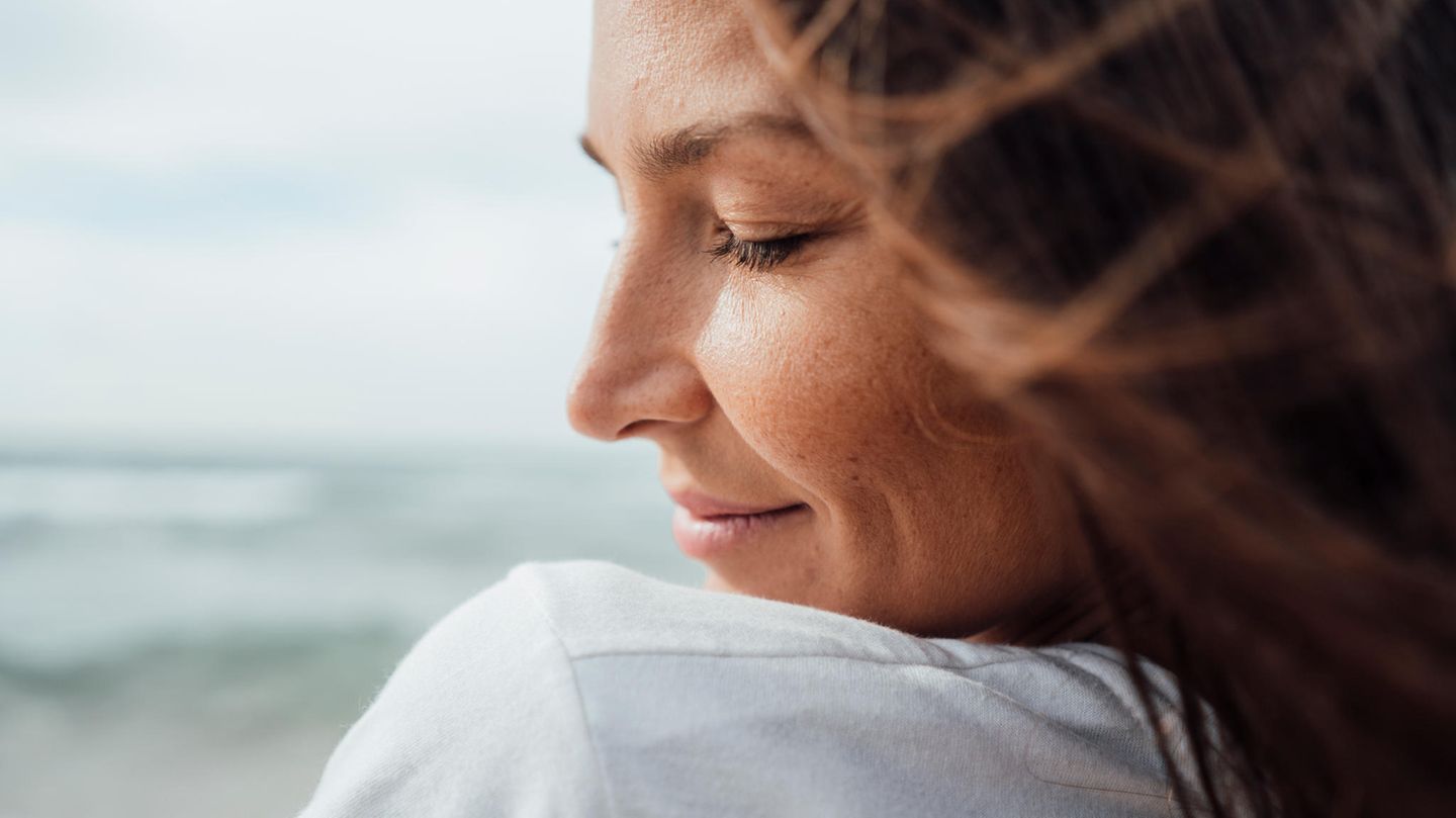 Eine Frau steht am Meer, hat die Augen geschlossen und lächelt