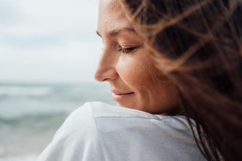 Eine Frau steht am Meer, hat die Augen geschlossen und lächelt