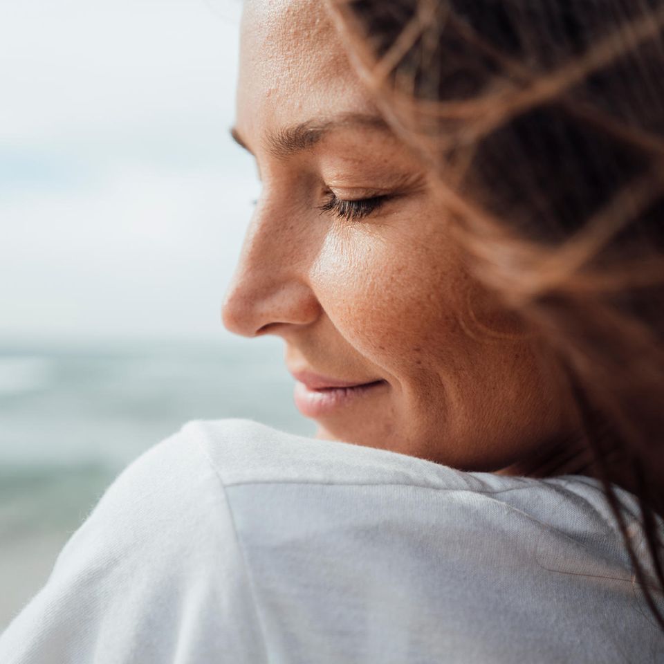 Eine Frau steht am Meer, hat die Augen geschlossen und lächelt