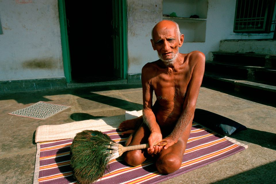 Ein älterer, nackter Mann sitzt auf einer dünnen Decke auf dem Boden. In seiner Hand hält er einen Wedel aus Pfauenfedern