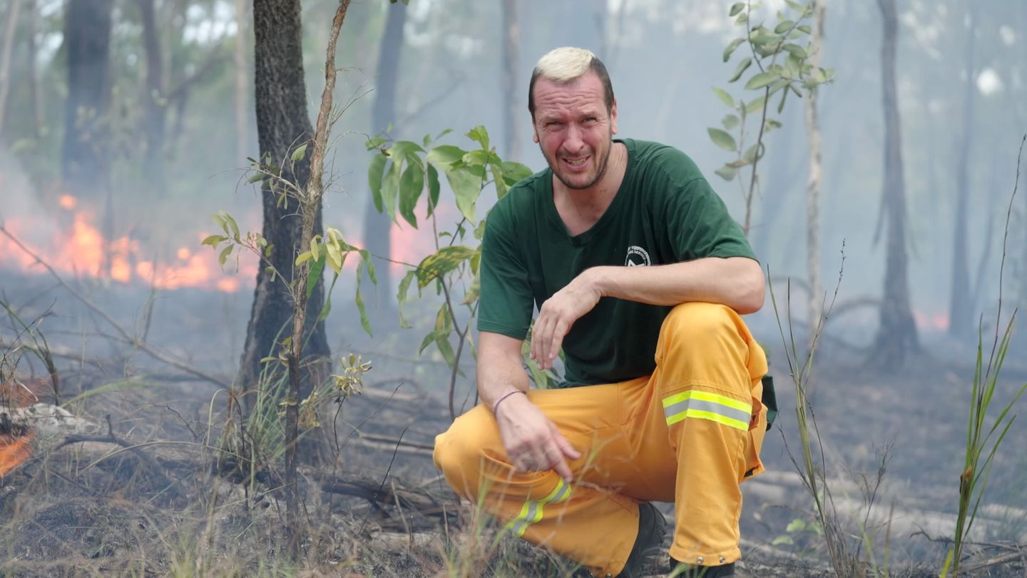 Pascal Hens kniet in einem Wald, hinter ihm ist Feuer zu sehen