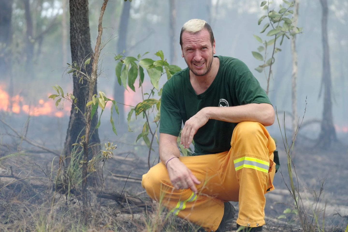 Pascal Hens kniet in einem Wald, hinter ihm ist Feuer zu sehen