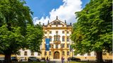 Gelb-weißes Schloss mit grünen Bäumen davor bei Sonnenschein
