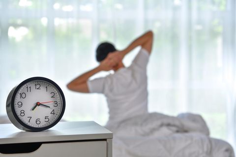 Ein Mann sitzt im Bett und streckt seine Arme – Lerchen und Eulen sind unterschiedlich früh wach bzw. müde