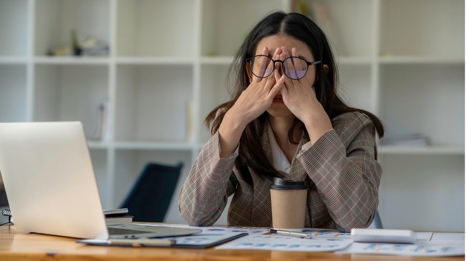 Eine Frau sitzt an einem Schreibtisch und hält sich erschöpft die Hände vor die Augen