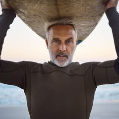 Ein älterer Mann stützt ein Surfbrett mit dem Kopf