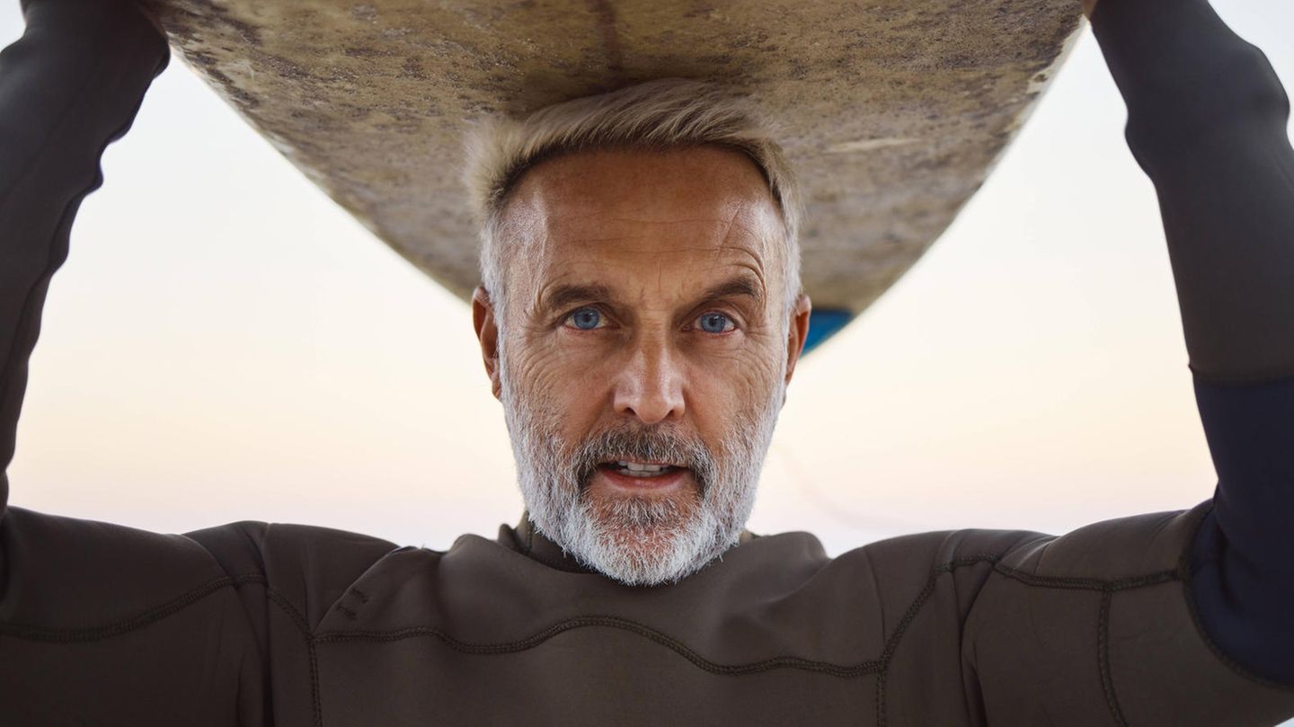 Ein älterer Mann stützt ein Surfbrett mit dem Kopf