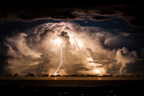 Große Gewitterwolke mit zwei herabzuckenden Blitzen über einer nächtlichen Bucht.