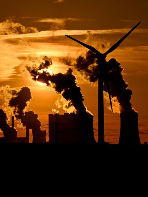 Abgase ausstoßende Schornsteine des Kohlekraftwerks Neurath, Strommasten und ein Windrad während des Sonnenuntergangs.