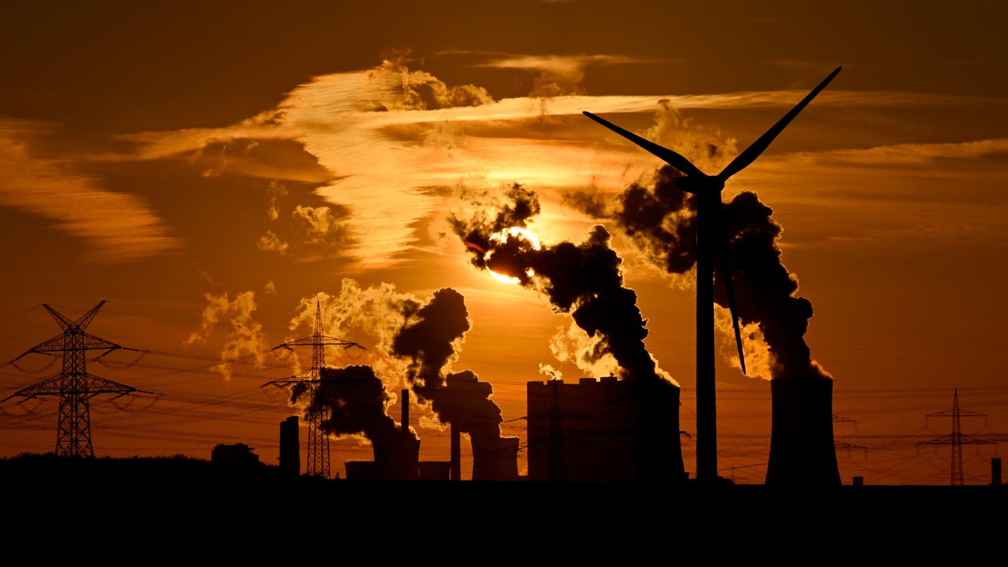 Abgase ausstoßende Schornsteine des Kohlekraftwerks Neurath, Strommasten und ein Windrad während des Sonnenuntergangs.