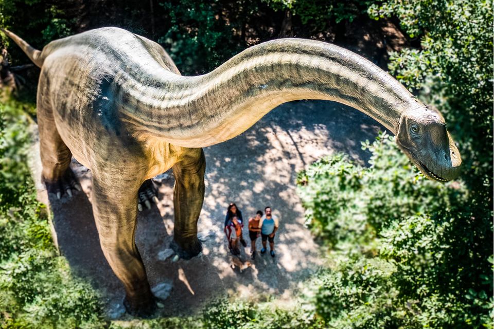 Luftaufnahme: Ein riesiges Dinosauriermodell neben einer winzig wirkenden Gruppe von Menschen