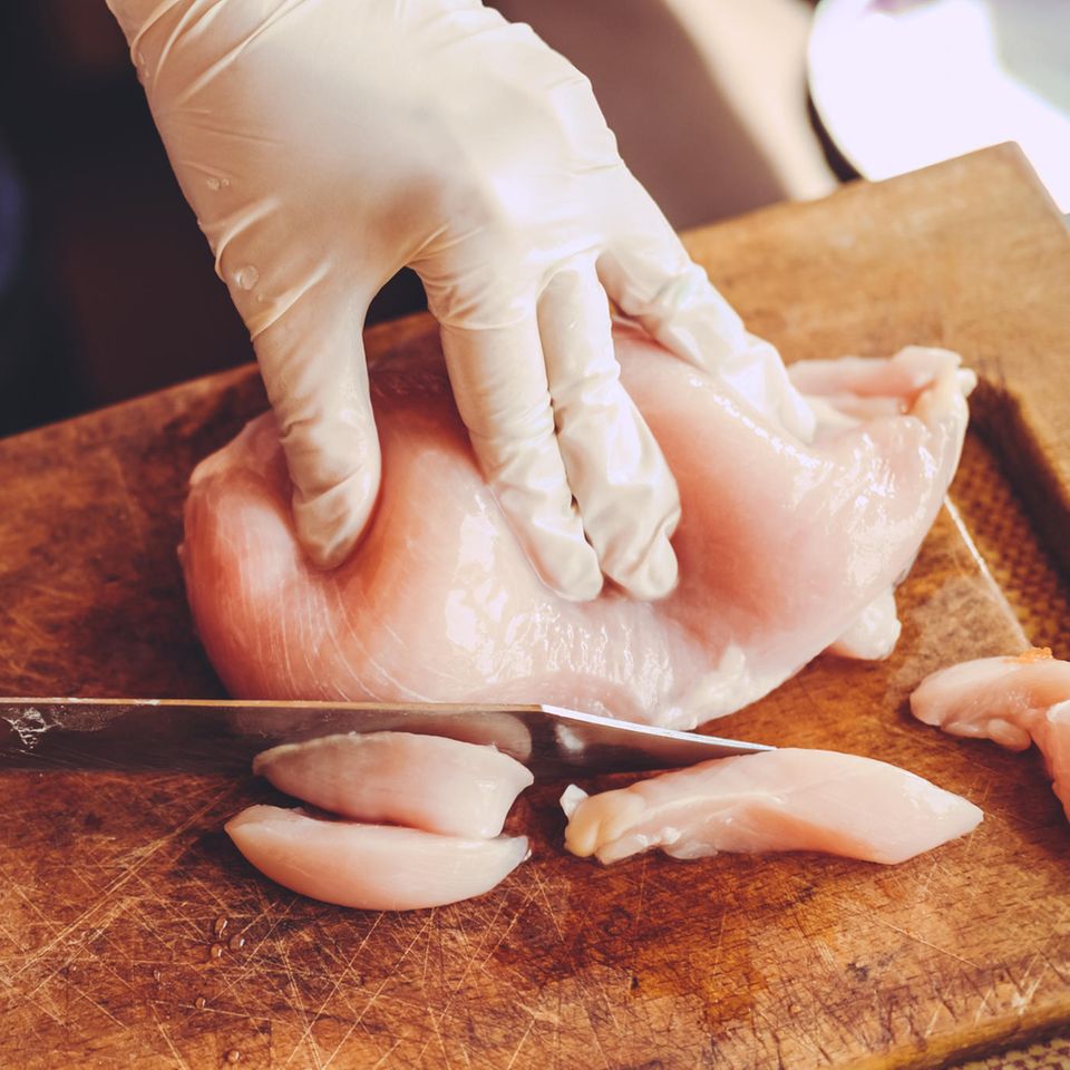 Rohes Hühnchenfleisch wird auf einem Holzbrett von einer Person mit Handschuhen kleingeschnitten Rohes Hühnchenfleisch wird auf einem Holzbrett von einer Person mit Handschuhen kleingeschnitten