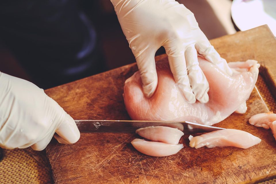 Rohes Hühnchenfleisch wird auf einem Holzbrett von einer Person mit Handschuhen kleingeschnitten  1.5004