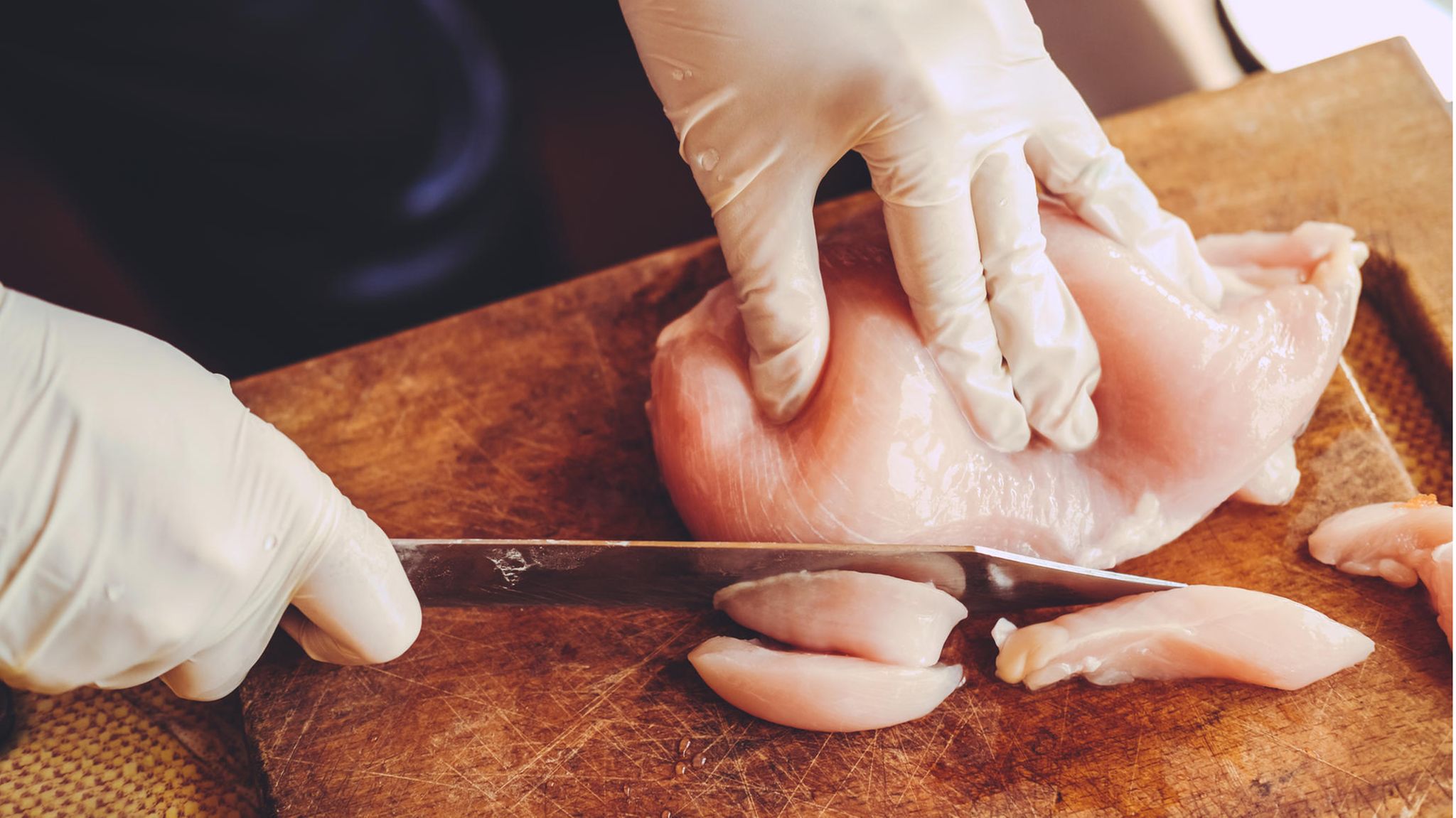 Rohes Hühnchenfleisch wird auf einem Holzbrett von einer Person mit Handschuhen kleingeschnitten