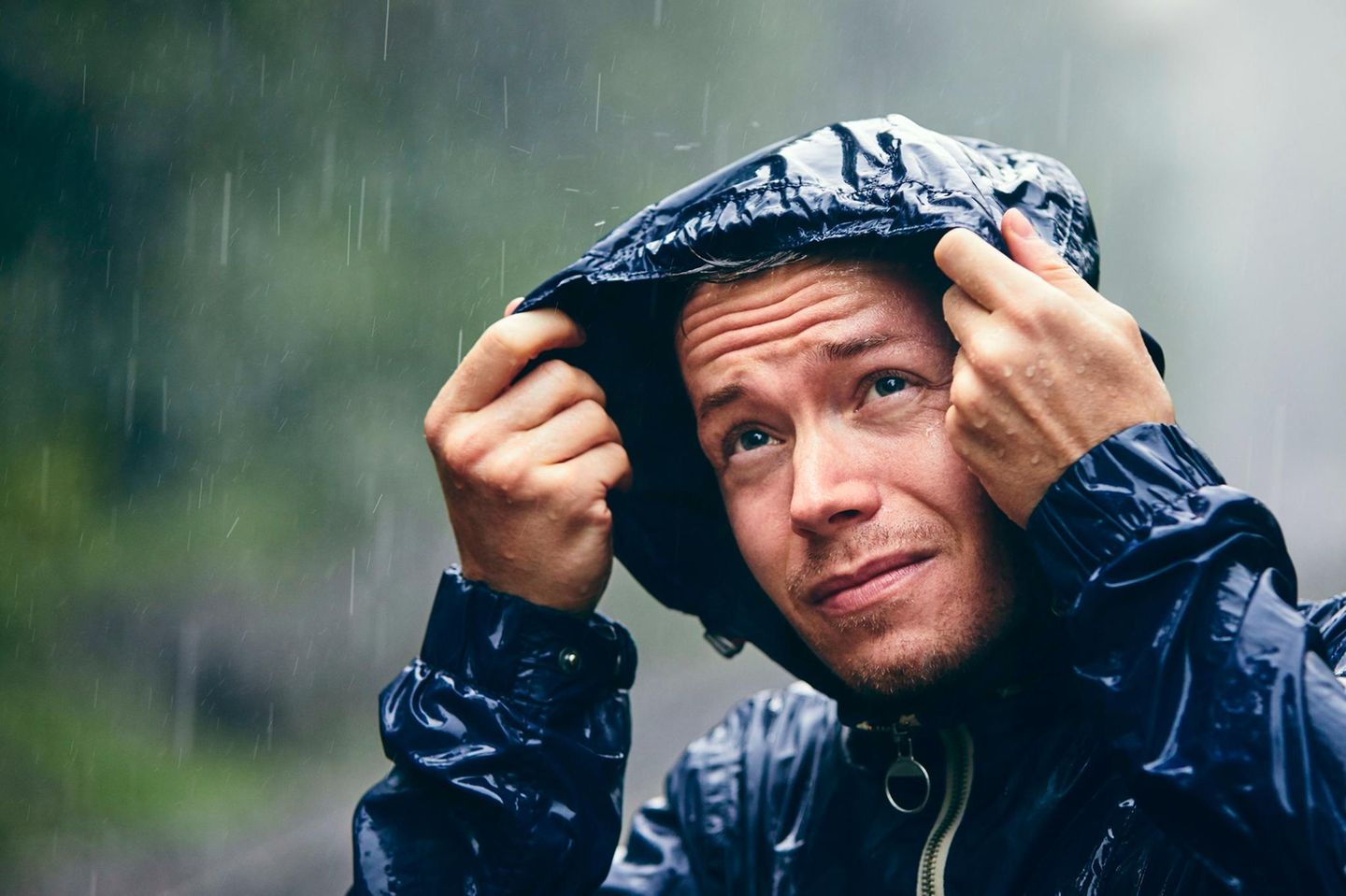 Ein Mann steht im Regen und schaut kritisch unter seinem Regencape