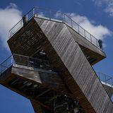 Der Turm ist aus Holz, die oberste Plattform schützt eine Reling aus Eisen. Der Himmel ist sommerlich weißblau.