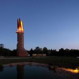 Der Turm steht in einer Abendstimmung am Waldrand, seine Spitze besteht aus vier Teilen, die beleuchtet sind.