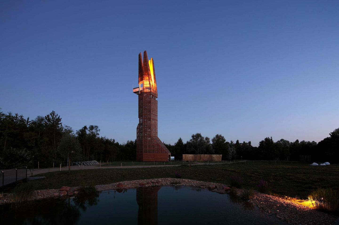 Der Turm steht in einer Abendstimmung am Waldrand, seine Spitze besteht aus vier Teilen, die beleuchtet sind.