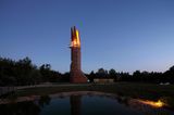 Der Turm steht in einer Abendstimmung am Waldrand, seine Spitze besteht aus vier Teilen, die beleuchtet sind.