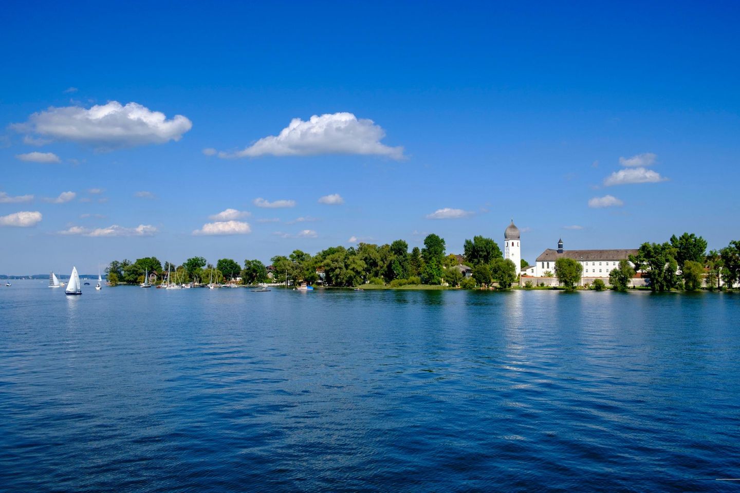 Abtei Frauenwörth, auf der Fraueninsel im Chiemsee Das weiße Kloster mit Zwiebelturm zwischen den Bäumen auf einer Insel in einem sehr blauen See.