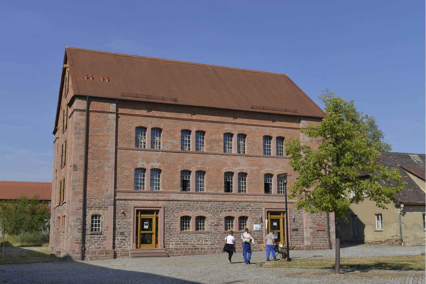 Kloster Helfta, bei Eisleben in Sachsen-Anhalt Ein großes Haus aus rotbraunen Ziegelsteinen mit Steinfundament. Drei Menschen gehen über einen Platz davor.