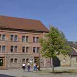 Ein großes Haus aus rotbraunen Ziegelsteinen mit Steinfundament. Drei Menschen gehen über einen Platz davor.