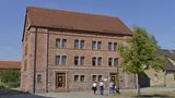 Ein großes Haus aus rotbraunen Ziegelsteinen mit Steinfundament. Drei Menschen gehen über einen Platz davor.