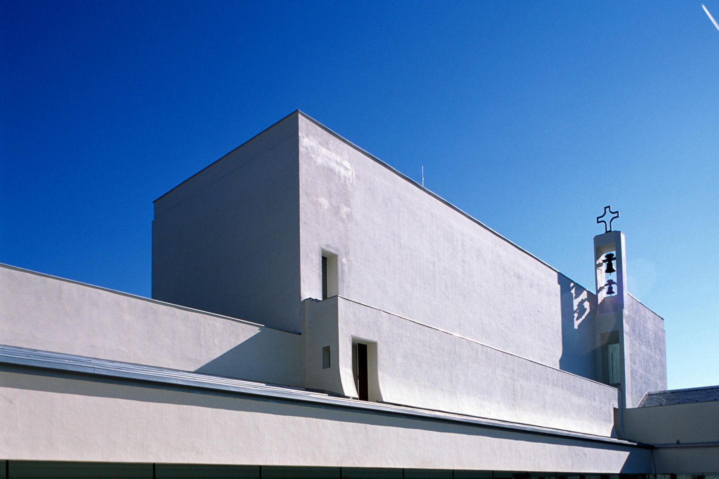 Ein Gebäude wie eine weiße Kiste, man sieht einen kleinen Glockturm mit einem Kreuz.