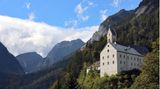 Großes, helles Haus mit einem burgähnlichen Turm am bewaldeten Hang unterhalb der Karwendelfelsen. Himmel mit ein paar Wolken.