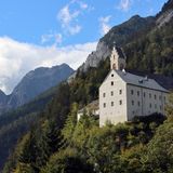 Großes, helles Haus mit einem burgähnlichen Turm am bewaldeten Hang unterhalb der Karwendelfelsen. Himmel mit ein paar Wolken.