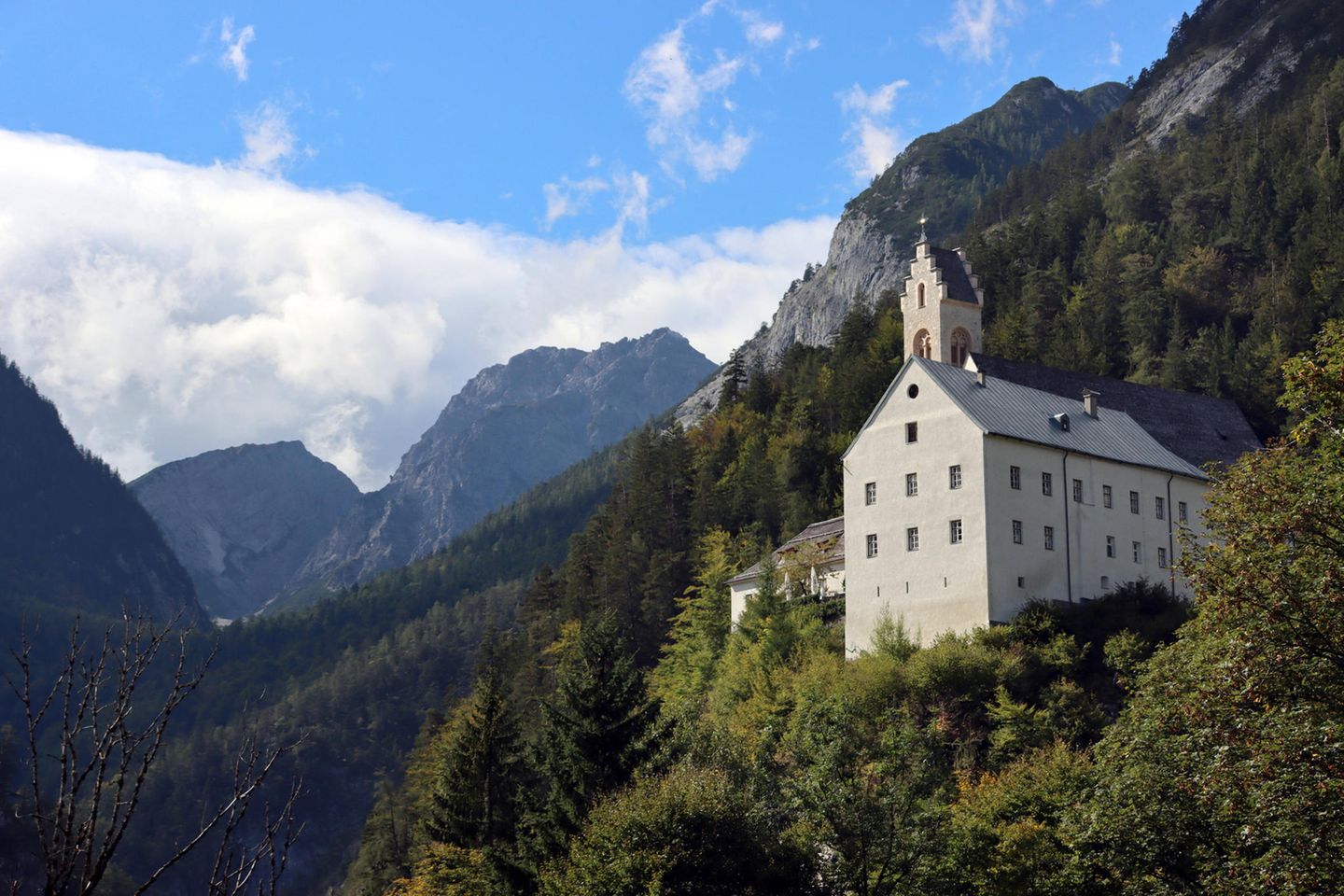 Großes, helles Haus mit einem burgähnlichen Turm am bewaldeten Hang unterhalb der Karwendelfelsen. Himmel mit ein paar Wolken.