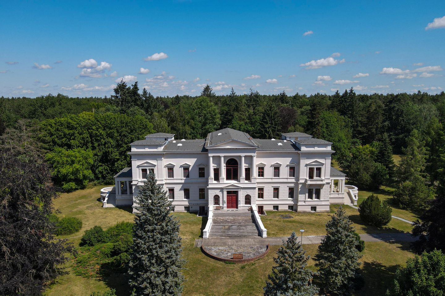 Weißes Schloss, eher Gutshaus mit Freitreppe und Wiese, dahinter Laub- und Nadelbäume.