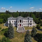 Weißes Schloss, eher Gutshaus mit Freitreppe und Wiese, dahinter Laub- und Nadelbäume.