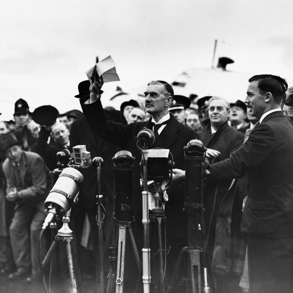 Der britische Premier Neville Chamberlain steht vor mehreren Mikrofonen.