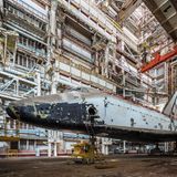 Eine rostende Raumfähre von der Seite fotografiert,  über ihr in der Halle ein roter Schwebekran.