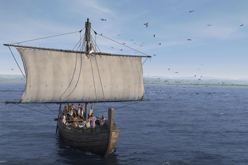 Ein offenes Holzschiff mit breitem Segel und elf Männern an Bord auf dem Meer, vor einem Küstenstreifen 1.5657