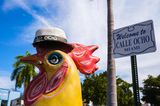 Eine Huhnfigur steht vor dem Straßenschild der Calle Ocho in Miami. Das Huhn hat einen Gut auf und eine Zigarre im Schnabel