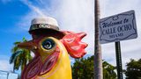 Eine Huhnfigur steht vor dem Straßenschild der Calle Ocho in Miami. Das Huhn hat einen Gut auf und eine Zigarre im Schnabel