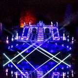 Ein Brunnen leuchtet festlich, die Bäume dahinter auch. Im Wasser spiegelt sich alles. Nachts.