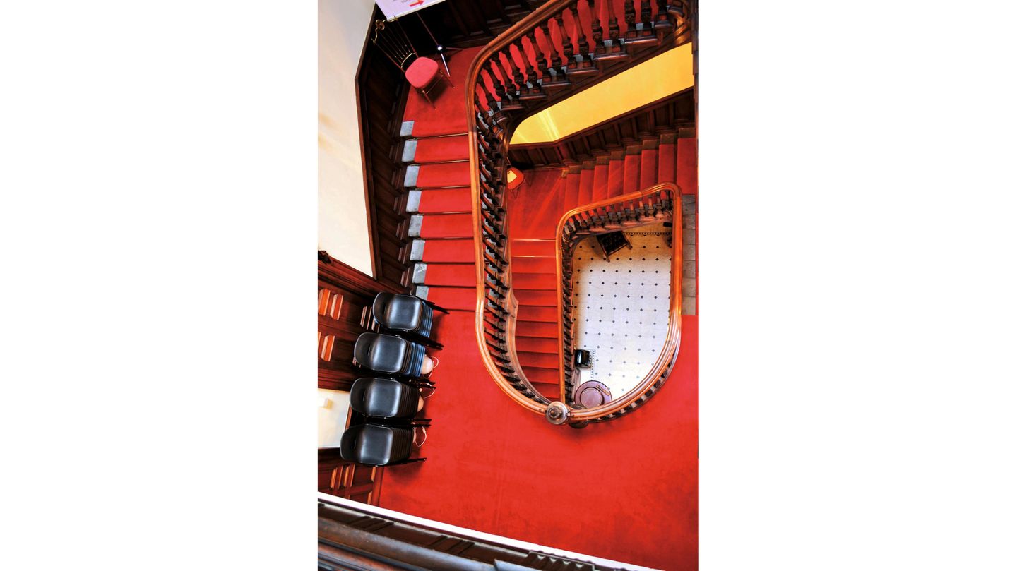 Blick von oben ins Treppenhaus, man sieht vor allem den roten Teppich auf der alten Treppe.