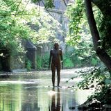 Eine mannshohe Eisenfigur steht auf einem Sockel im ruhigen Flüsschen, viel Grün am Ufer.