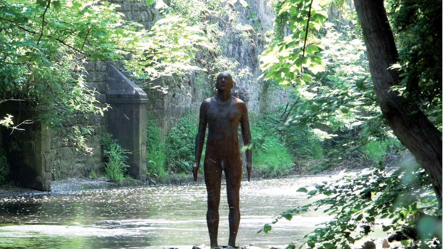 Eine mannshohe Eisenfigur steht auf einem Sockel im ruhigen Flüsschen, viel Grün am Ufer.