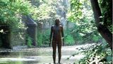 Eine mannshohe Eisenfigur steht auf einem Sockel im ruhigen Flüsschen, viel Grün am Ufer.