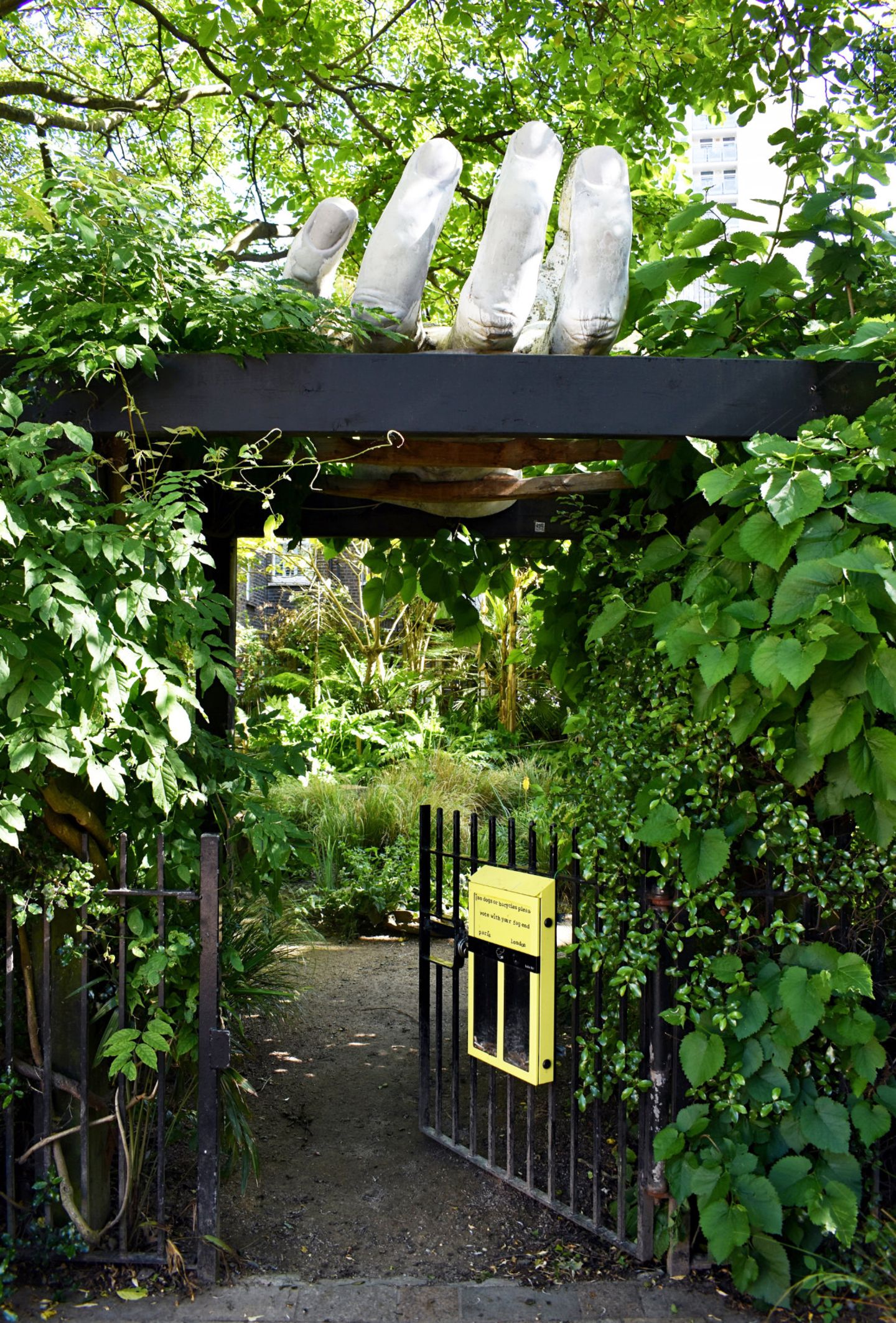 Alles Handarbeit: der Bonnington Square Pleasure Garden Ein dicht eingewachsenes Gartentor mit einer großen Handskulptur, die darüber schwebt.