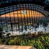 Großes, bodentiefes, halbrundes Fenster überspannt einen Platz mit Tischen und Pflanzen. Man sieht die Skyline Londons