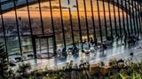 Großes, bodentiefes, halbrundes Fenster überspannt einen Platz mit Tischen und Pflanzen. Man sieht die Skyline Londons