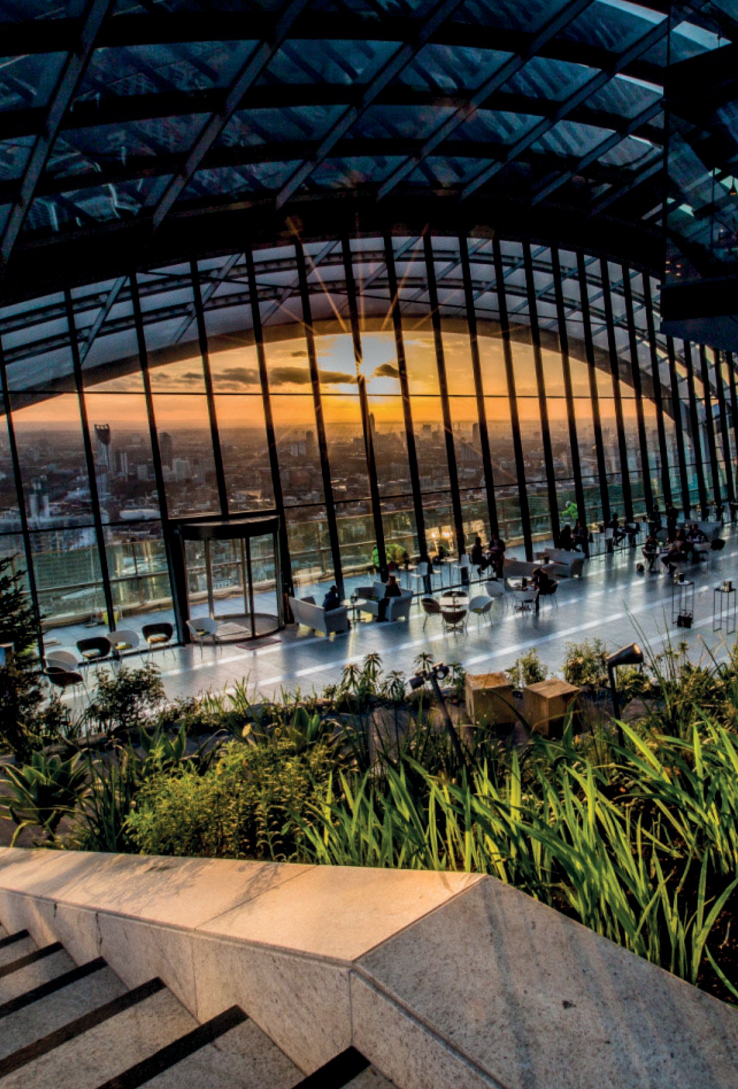 Ganz weit oben: Sky Garden Großes, bodentiefes, halbrundes Fenster überspannt einen Platz mit Tischen und Pflanzen. Man sieht die Skyline Londons